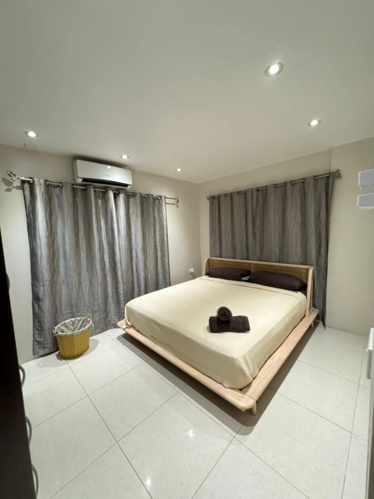 Quarto moderno com uma cama grande, cortinas cinzas, um pequeno cesto e um aparelho de ar condicionado.