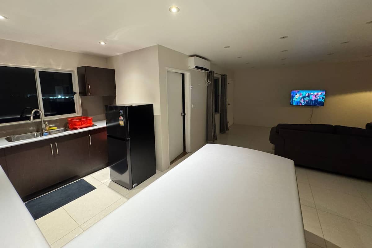 A modern kitchen with a refrigerator, sink, and window. There is a TV mounted on the wall and a dark-colored sofa in the living area.