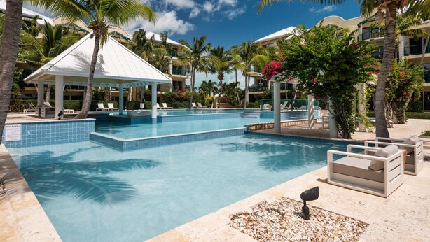 Área de piscina em resort, com águas azul-claras, espreguiçadeiras, um gazebo e palmeiras.