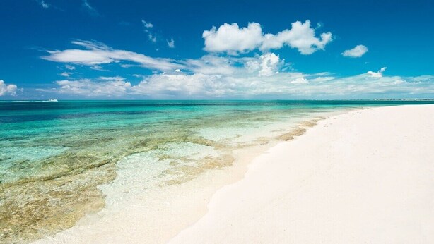 Ein Strand mit klarem, türkisblauem Wasser und weißem Sand unter blauem Himmel mit Wolken.
