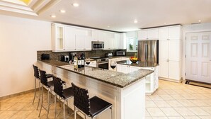 Eine moderne Küche mit weißen Schränken, Granit-Arbeitsplatten und Edelstahlgeräten. Es gibt drei Barhocker und eine große Kochinsel mit Spüle.