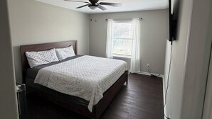 A bedroom with a bed, a ceiling fan, a window with curtains, and a wall-mounted TV. - Comfortable and safe family lodging for everyone close to the medical center.  (Houston)