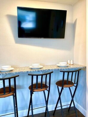 A kitchen with a TV mounted on the wall, a granite countertop, and three bar stools.