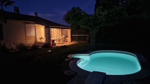 A backyard with a swimming pool, a patio with outdoor furniture, and a house with a lit interior.