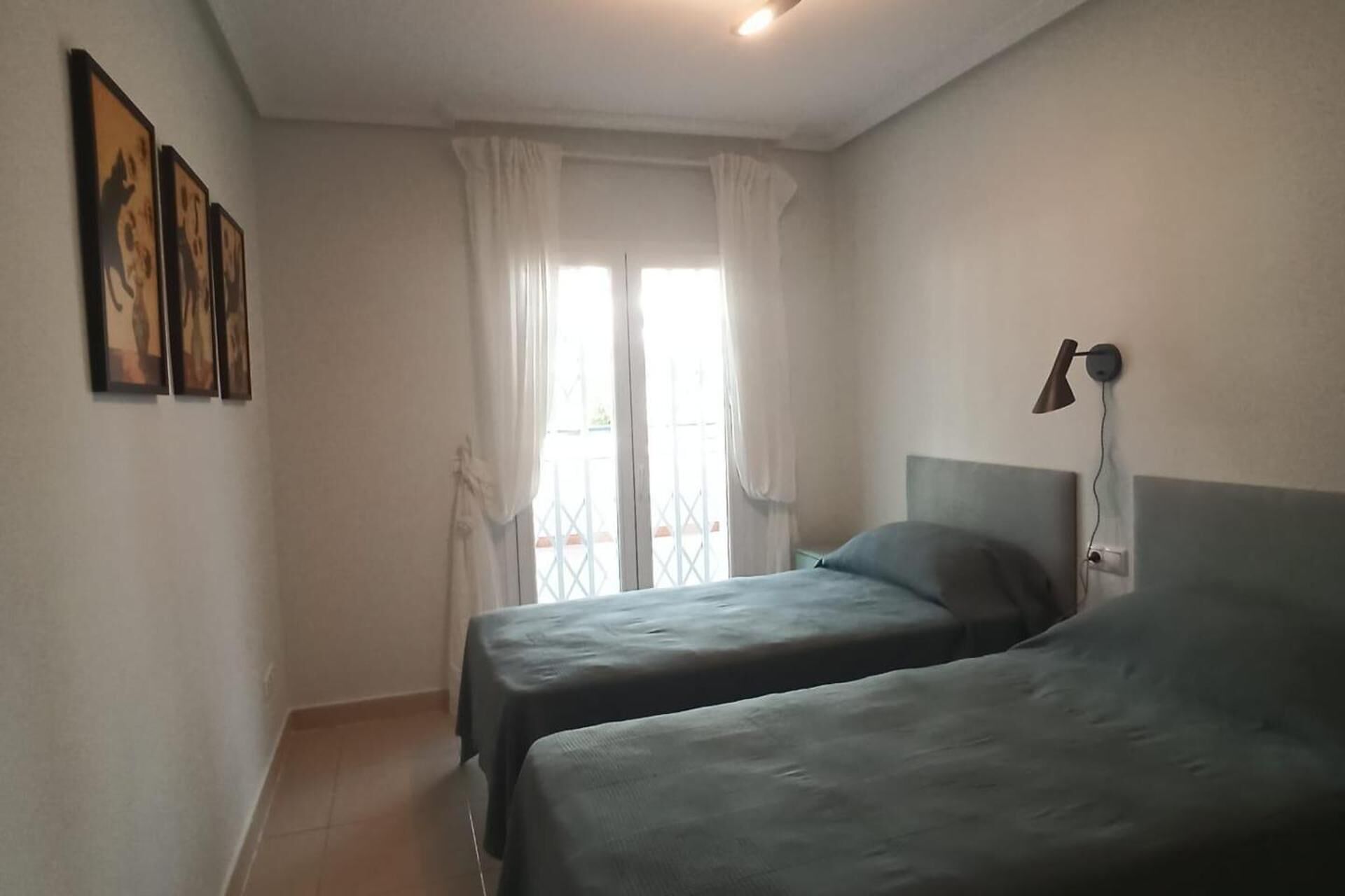 Una habitación con dos camas, una ventana con cortinas blancas, una lámpara de pared y cuadros enmarcados.
