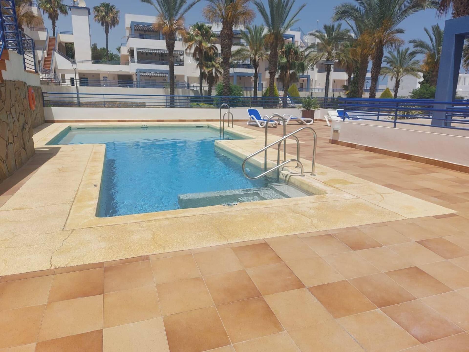 Piscina exterior con borde de piedra, escalera metálica y sillas reclinables azules.