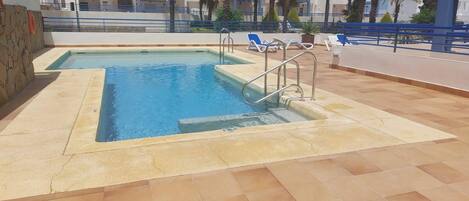 Piscina exterior con borde de piedra, escalera metálica y sillas reclinables azules.