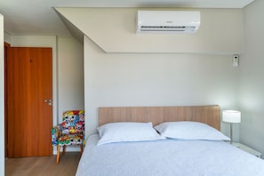 Een slaapkamer met een bed, een houten hoofdbord, een kleurrijke stoel, een witte lamp en een airconditioning.