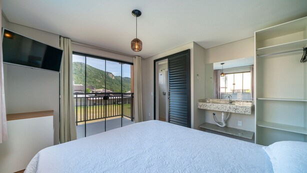 Een slaapkamer met een groot bed, een televisie, een balkon met uitzicht op de heuvels en een badkamer met een wastafel en spiegel.