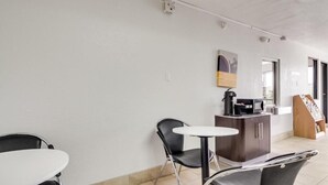 A small, clean room with a white table, two black chairs, a small cabinet with a coffee machine, and a wooden rack with magazines. - AVERA SUITES (Muskogee)