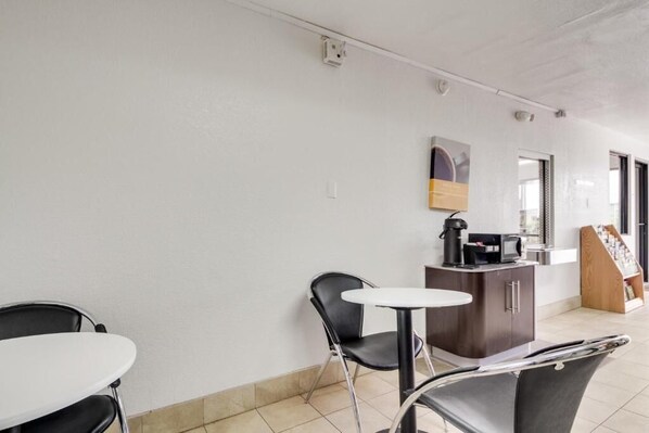 Une petite pièce propre, avec une table blanche, deux chaises noires, un petit meuble contenant une machine à café et un présentoir en bois avec des magazines.