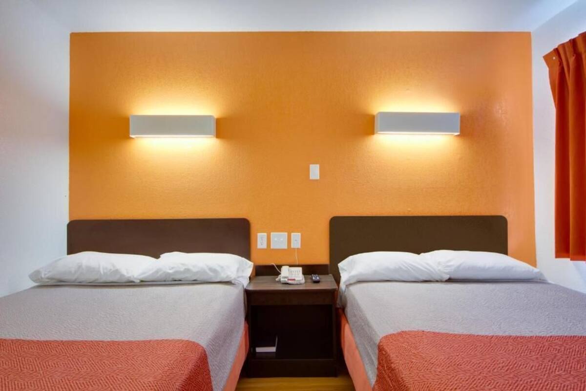 Ein Hotelzimmer mit zwei Einzelbetten, einem Nachttisch mit Telefon und Fernbedienung sowie einer wandmontierten Lampe.