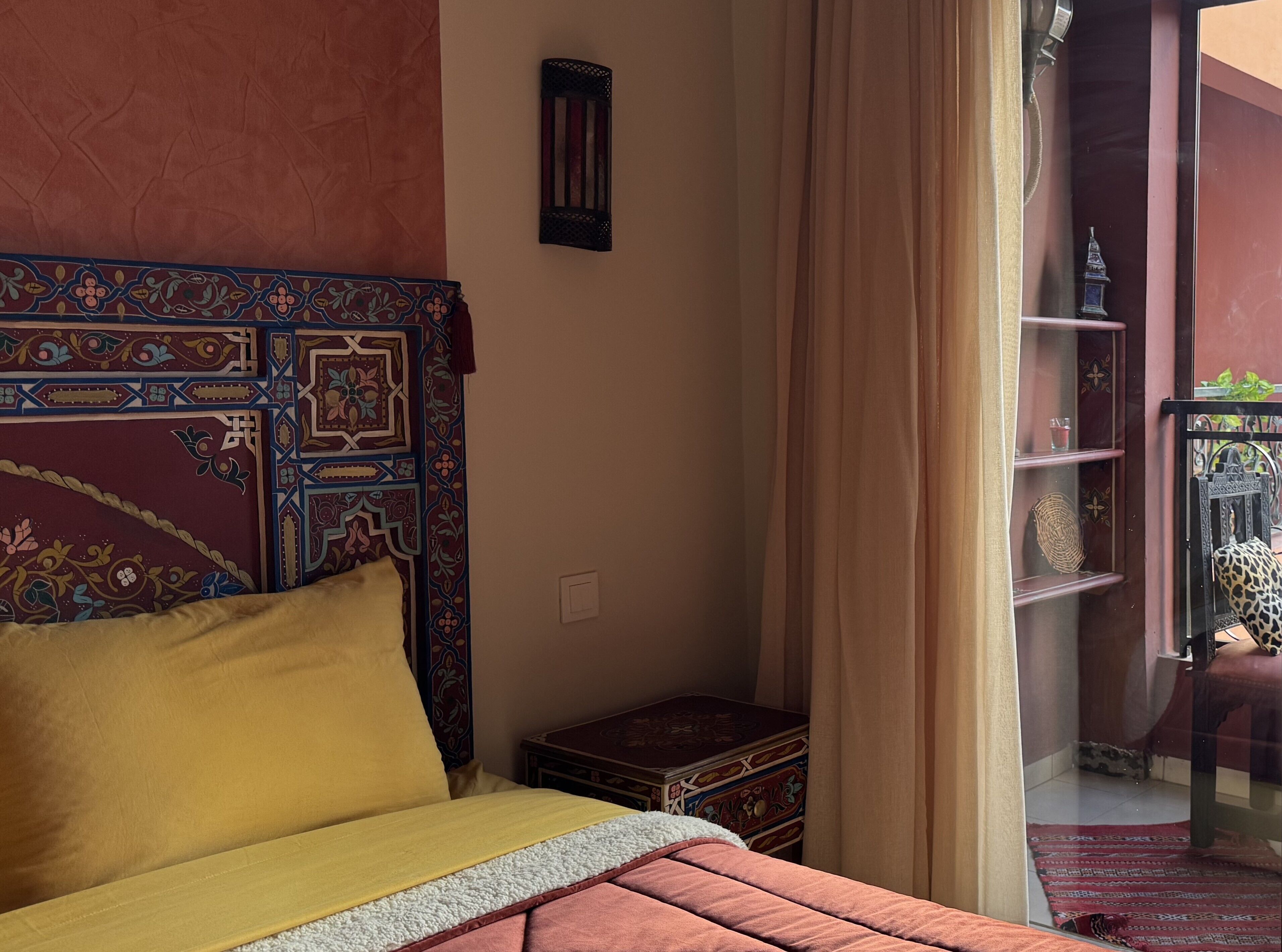 A bed with a patterned headboard, a small wooden chest, and a view of a balcony with a chair and a plant.