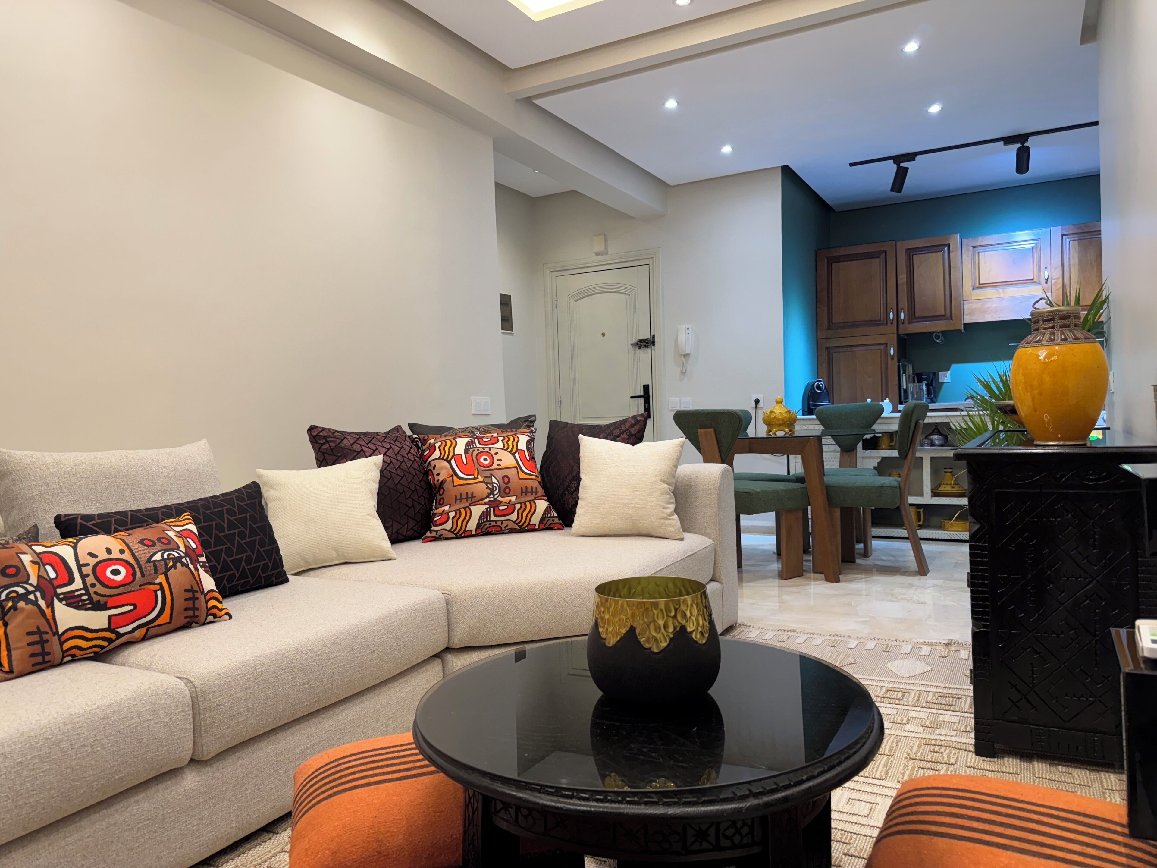 A modern living room with a beige sofa, colorful cushions, a glass coffee table, and a kitchen area in the background.