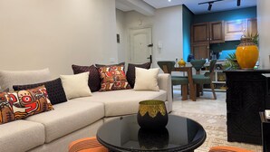 Ein modernes Wohnzimmer mit einem beigen Sofa, farbenfrohen Kissen, einem Glastisch und einer Küchenzeile im Hintergrund.