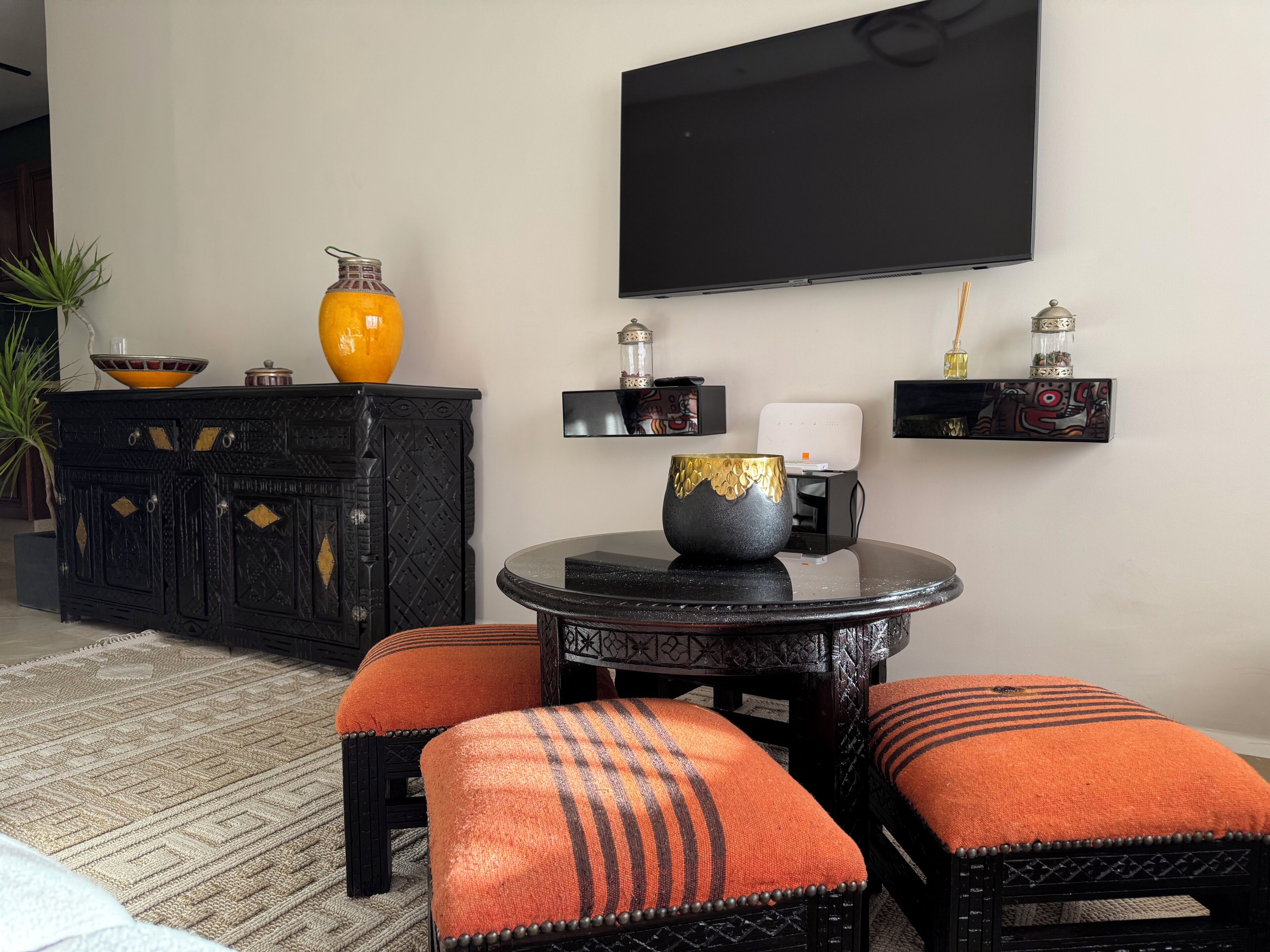 Een woonkamer met een zwart kastje, oranje kussens, een ronde tafel en een flatscreen televisie aan de muur.