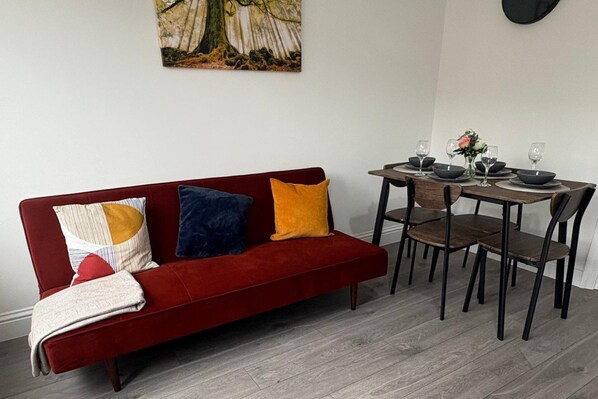 Un sofá rojo con cojines estampados, una mesa de comedor preparada con platos y vasos, y un cuadro en la pared.