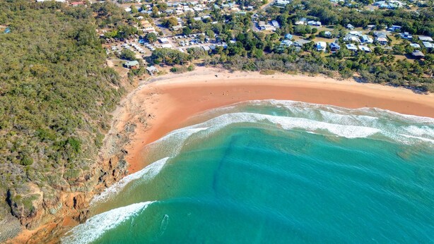 An aerial view of a coastal area with a sandy beach, clear turquoise waters, and a residential area with houses and greenery.