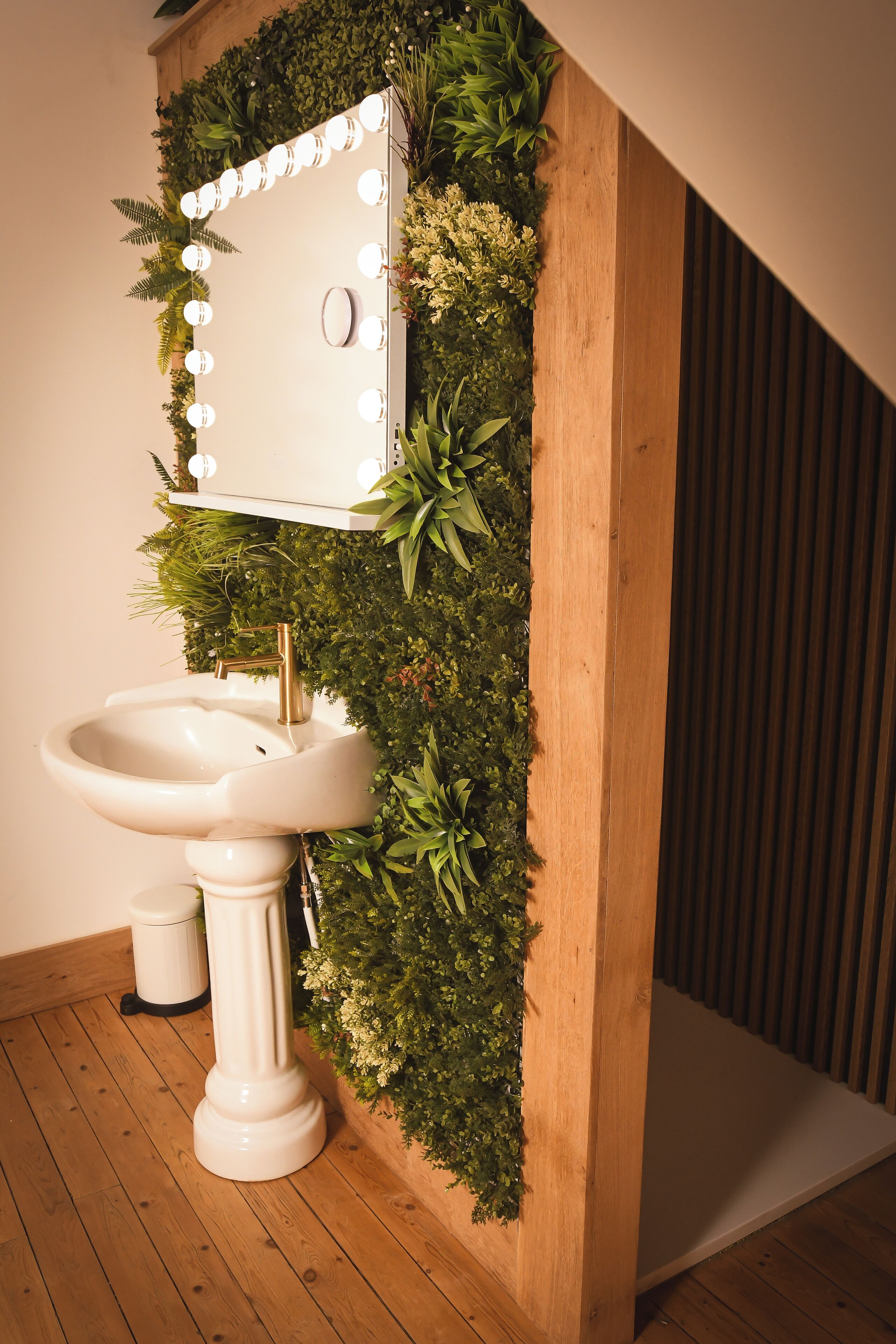 A bathroom with a white sink, a mirror with lights, and a wooden column with greenery.