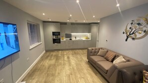 A modern living room with a brown leather sofa, a flat-screen TV mounted on the wall, and a kitchen area with white cabinets and a built-in oven. - Chic and spacious 2 bedroom, 2 bath Apartment (Epsom)