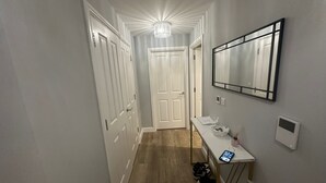 A narrow hallway with wooden flooring, white walls, and a ceiling light fixture. There is a door at the end of the hallway and a small table with a mirror and a phone on it. - Chic and spacious 2 bedroom, 2 bath Apartment (Epsom)