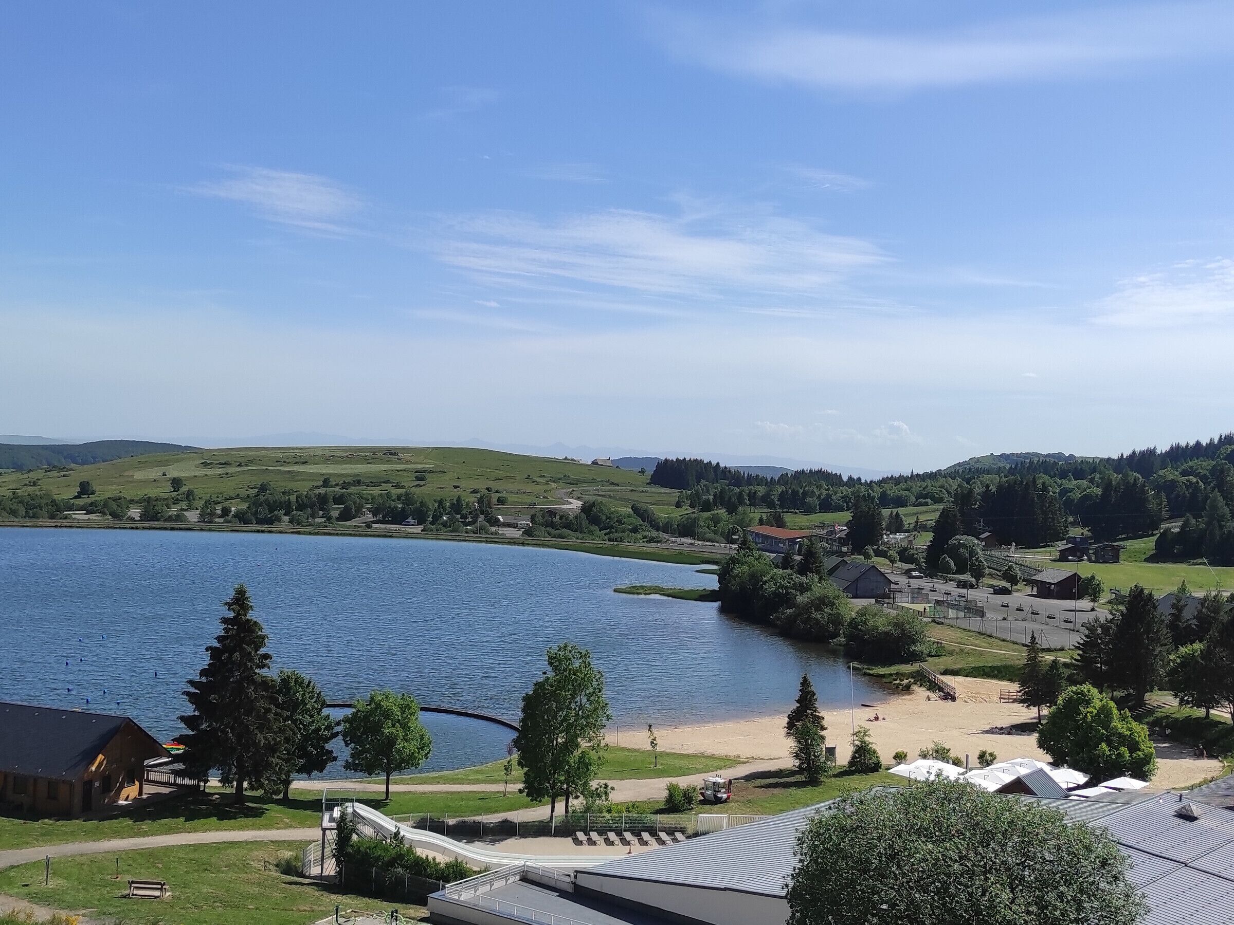 Vista de un lago con un pequeño muelle, una playa de arena y una colina al fondo.