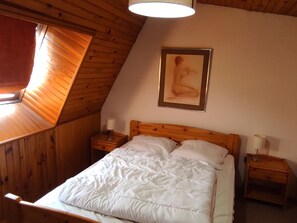 Une chambre avec un plafond en pente, des murs en bois, un lit avec une literie blanche, deux tables de chevet avec des lampes et une œuvre d’art encadrée accrochée au mur.