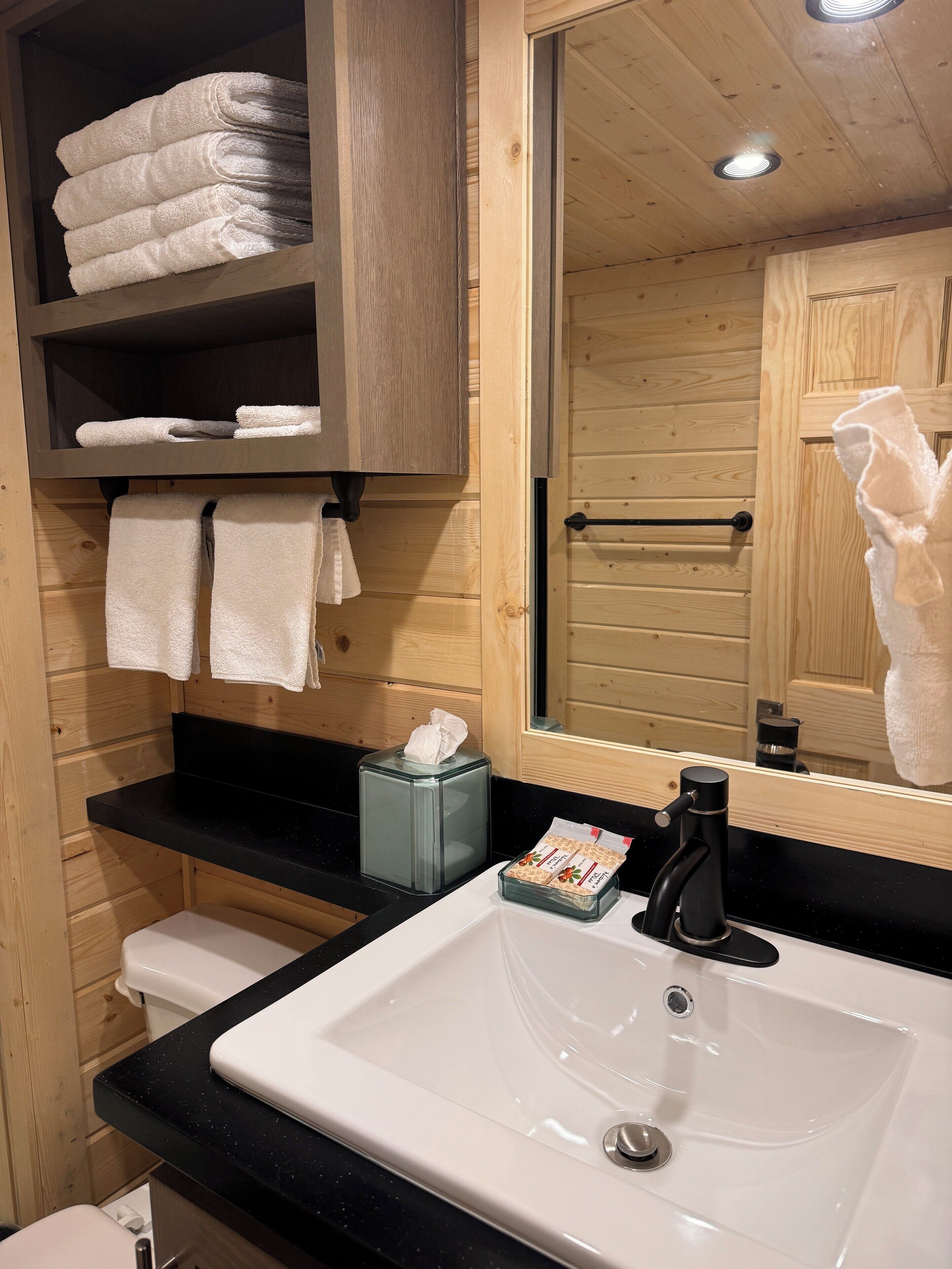 A bathroom with a wooden interior, a white sink, a black countertop, and a mirror. There are towels on a shelf and a tissue box on the counter.