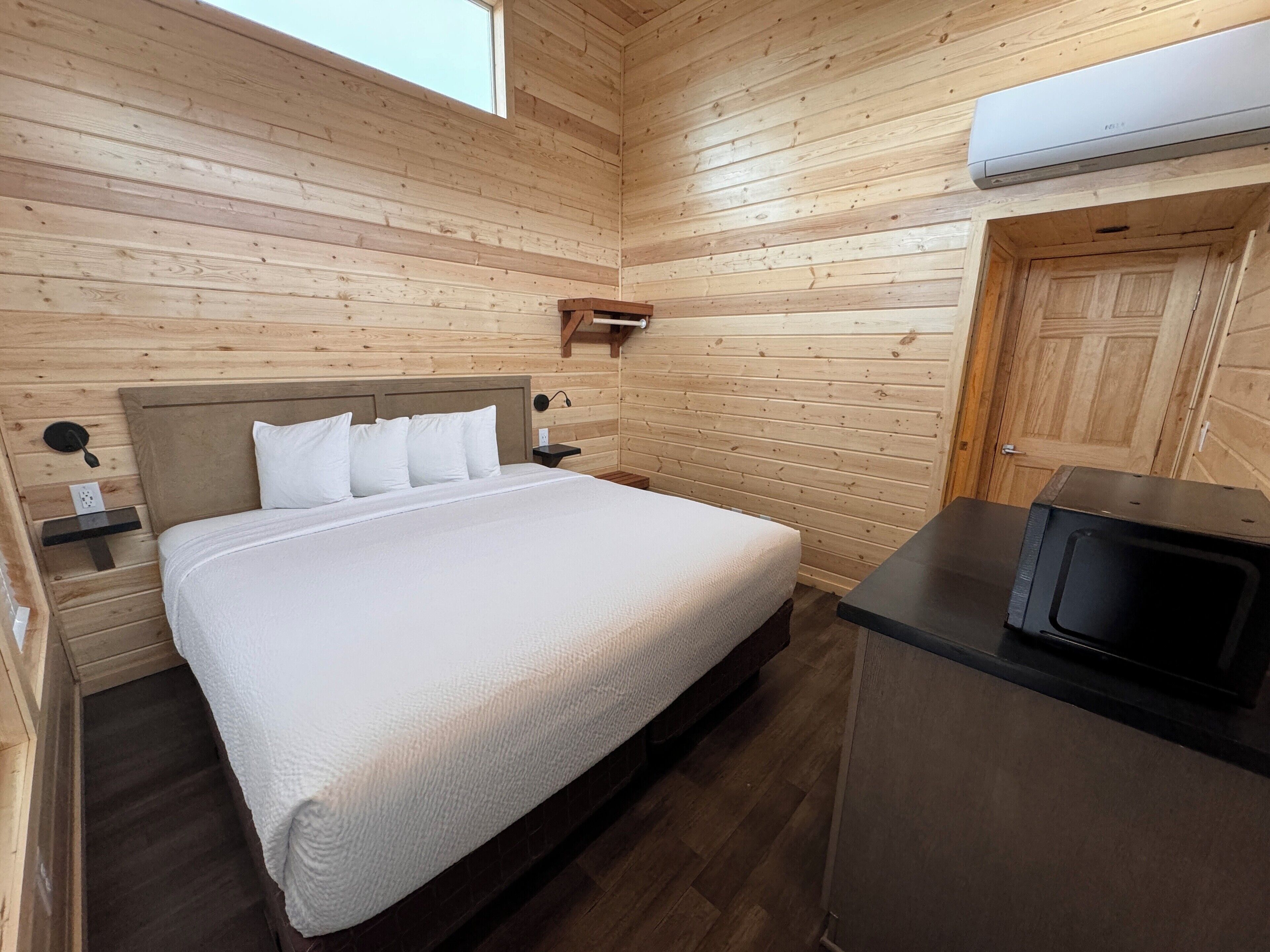A wooden cabin room with a bed, bedside tables, a microwave, and an air conditioning unit.
