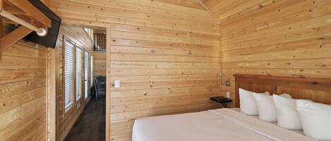 Interior de una cabaña de madera con una cama, un televisor montado en la pared y un pasillo con ventanas.