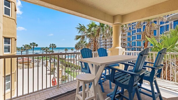 Ein Balkon mit weißem Tisch und blauen Stühlen, der Blick auf einen Strand und einen Poolbereich geht.