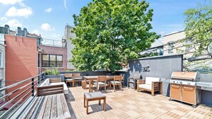 Un patio en la azotea con mobiliario de madera, una parrilla y un árbol grande que da sombra.