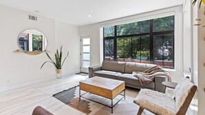 Una sala moderna con un sofá, una mesa de centro y un sillón. Hay un ventanal con vistas a árboles y un edificio.