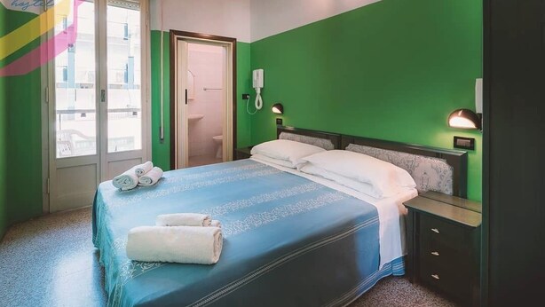 Une chambre d’hôtel comprenant un lit, une table de chevet, une commode, un téléphone fixé au mur et une fenêtre donnant sur l’extérieur.