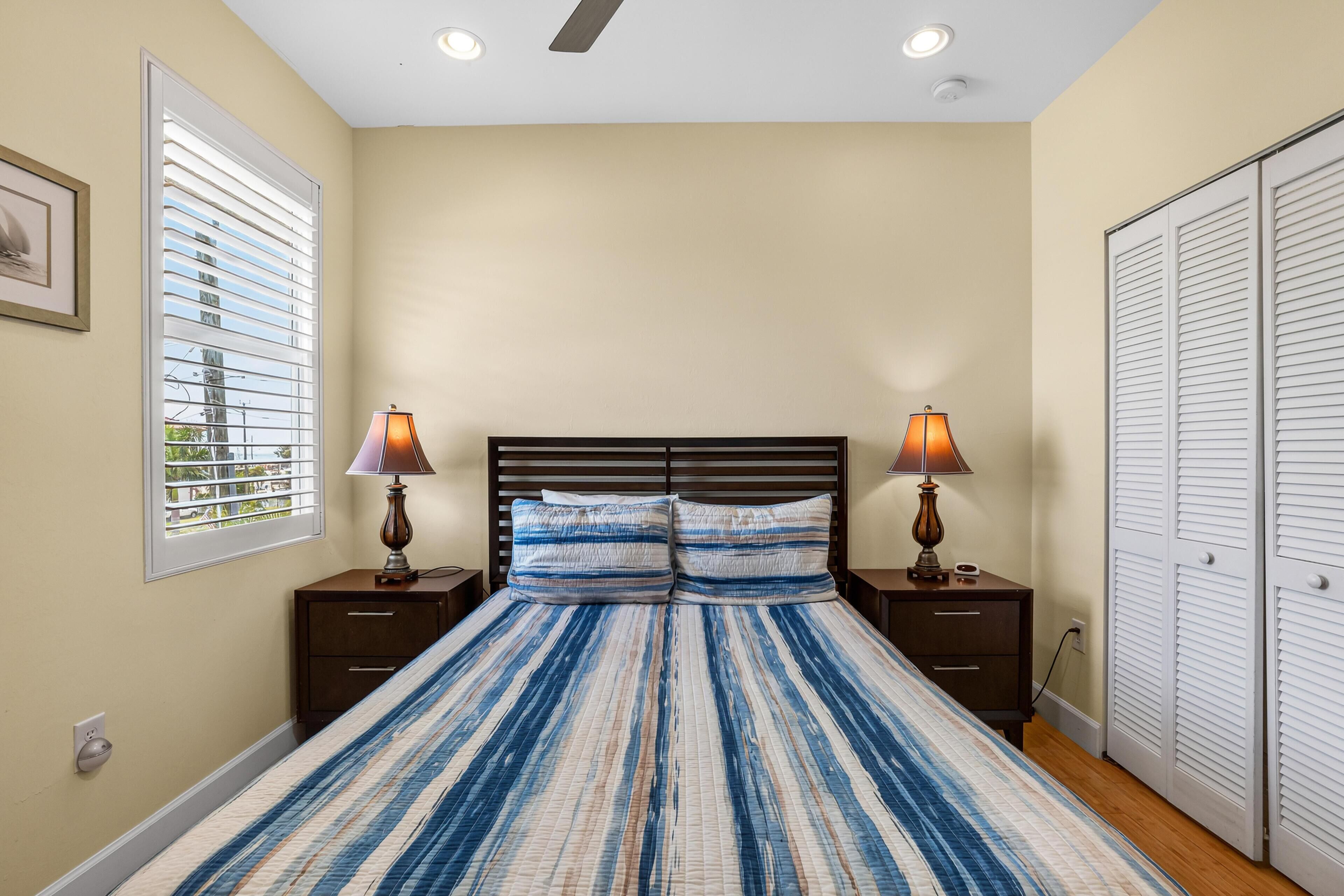 A bedroom with a bed, two bedside lamps, a nightstand, a window with blinds, and a closet.