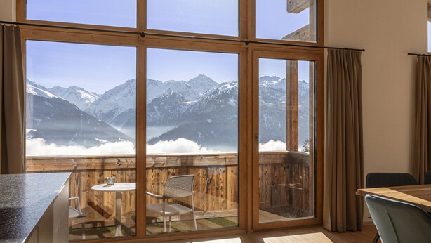 Ein Zimmer mit großen Fenstern und Bergblick, eine Holzterrasse mit Tisch und Stühlen sowie ein Blick zum Himmel.