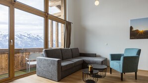 Een moderne woonkamer met een grijze hoekbank, een blauwe fauteuil en een kleine ronde tafel met een kaars. Grote ramen bieden uitzicht op besneeuwde bergen.