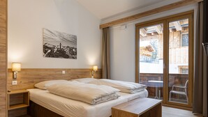 Slaapkamer met een houten hoofdeinde, twee nachtkastjes met lampen, een bed met witte lakens, een bankje aan het voeteneinde van het bed en een groot raam met uitzicht op een houten terras en zitplaatsen buiten.