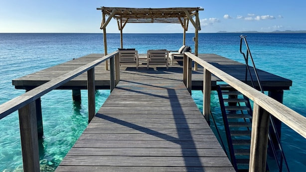 Un molo di legno che conduce a una spiaggia con acque blu cristalline e un gazebo in legno all'estremità.