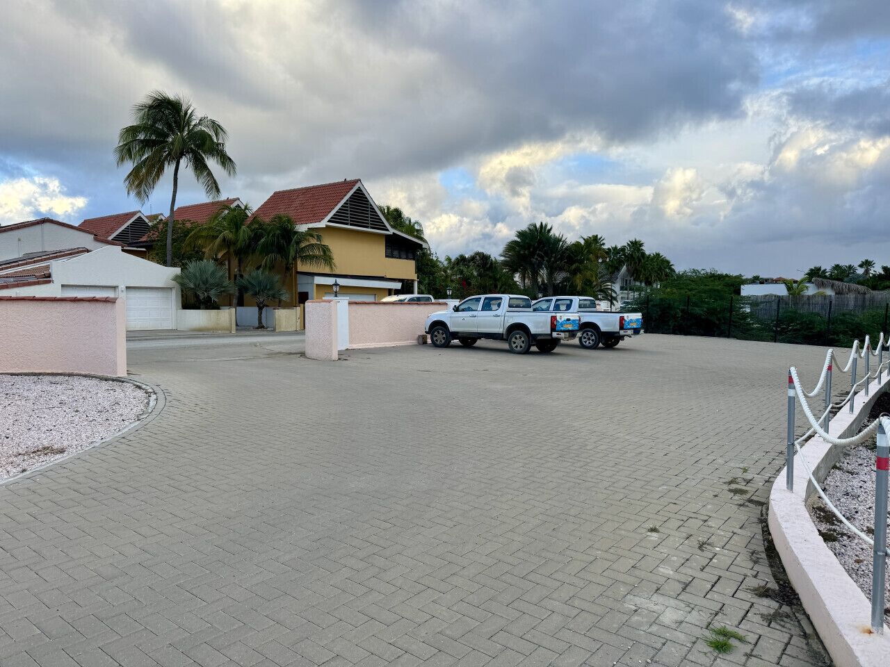 A paved driveway leading to a building with a red roof, surrounded by palm trees and a cloudy sky.