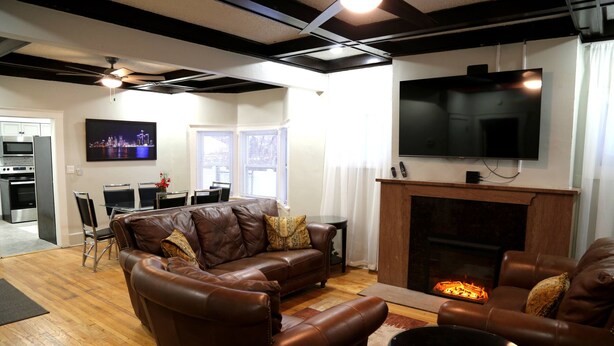 A modern living room with a fireplace, leather sofas, a flat-screen TV, and a dining area with a table and chairs. - Cozy 3-Bed Retreat with Games, Smart TVs & Fire Pit, 12 Mins to Downtown! (Detroit)