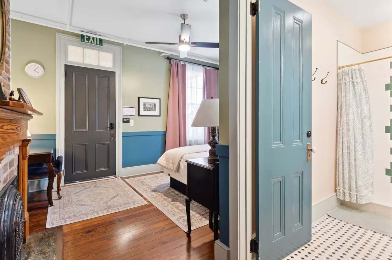 A room with a bed, a desk, a chair, a lamp, and a door with an exit sign.
