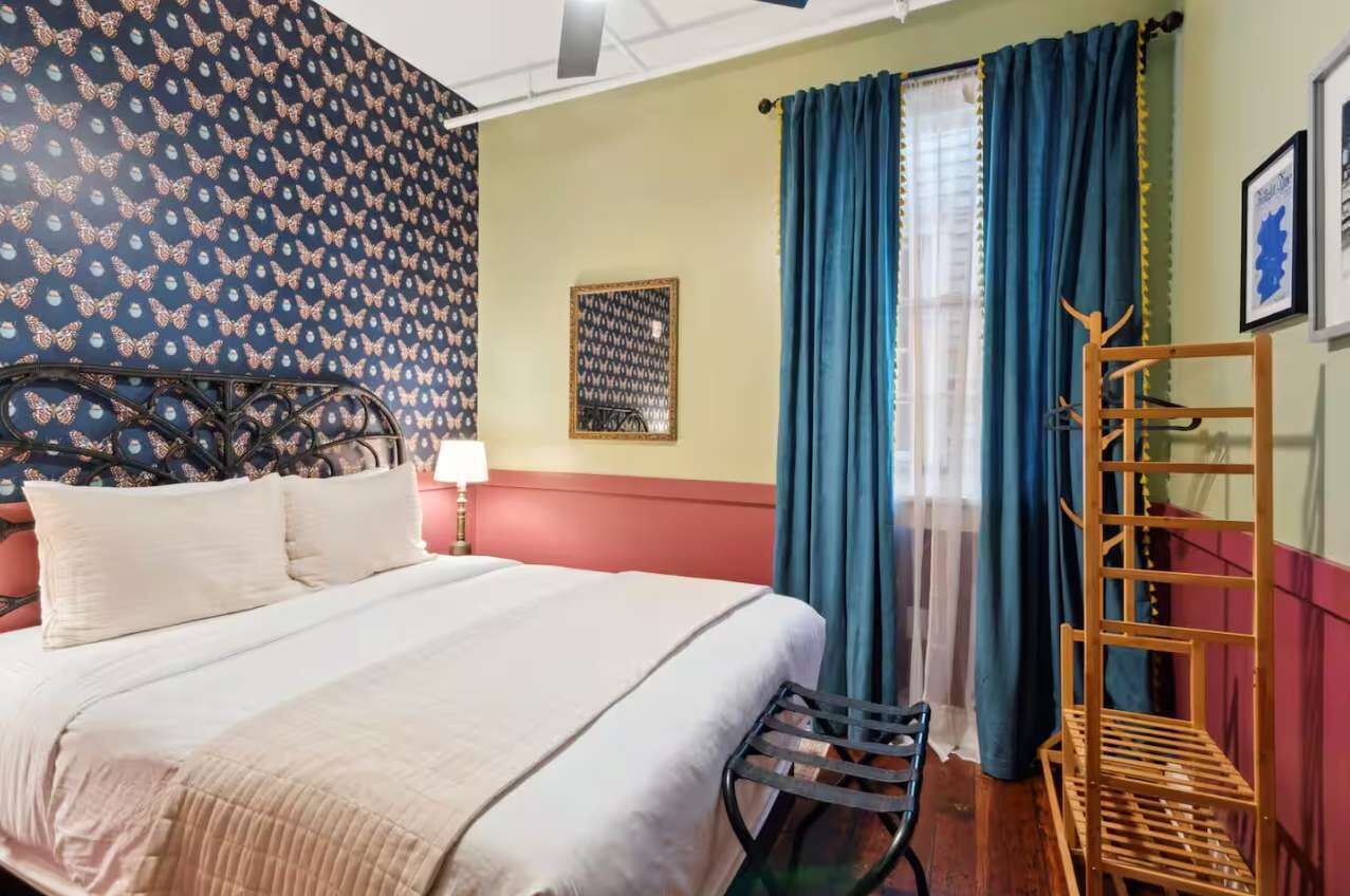 A hotel room with a bed, a chair, a wooden rack, a wall with a butterfly pattern, a framed picture, and a window with blue curtains.