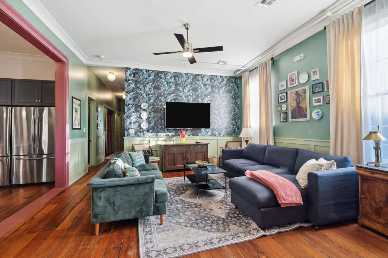 A living room with a sofa, armchair, and coffee table. There is a television mounted on the wall, a ceiling fan, and a kitchen area visible through an open door.