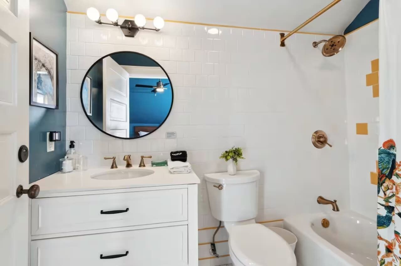 Une salle de bain moderne avec un lavabo blanc, des toilettes et une baignoire. Un miroir est placé au-dessus du lavabo, et on aperçoit une pomme de douche sur la droite.