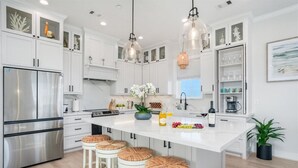 Una cocina moderna con muebles blancos, un refrigerador de acero inoxidable, una isla central con taburetes, iluminación colgante y una planta en maceta.