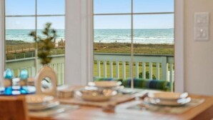 Una mesa de comedor con platos y vasos, un jarrón con flores y vistas al océano a través de amplias ventanas.