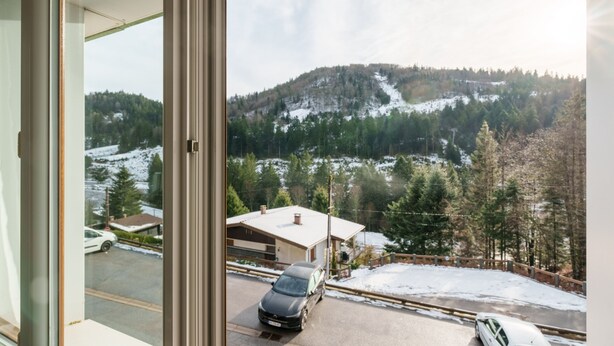 Une vue depuis une fenêtre donnant sur un paysage enneigé, avec une voiture garée dans l’allée et un bâtiment au premier plan.