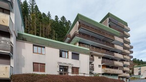 Un immeuble résidentiel à plusieurs étages, doté de balcons, d’un toit végétalisé et d’une haie devant l’entrée.