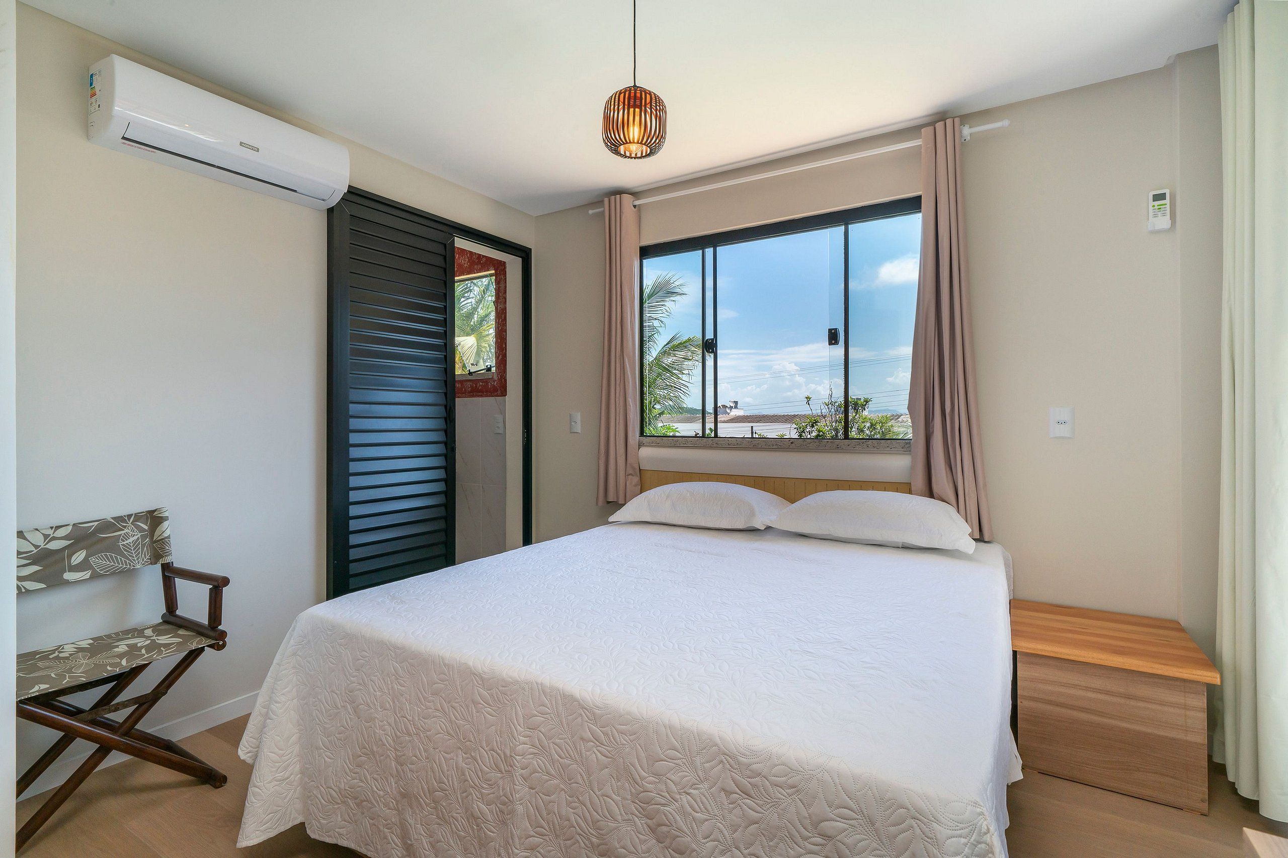 Un dormitorio con cama, una silla, un equipo de aire acondicionado, una ventana con cortinas y vistas al exterior.